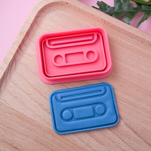May include: Two plastic cookie cutters shaped like cassette tapes. The top cutter is pink and the bottom cutter is blue.
