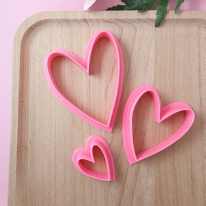 Op de afbeelding: Drie roze hartvormige koekjesvormen in verschillende maten op een houten oppervlak.
