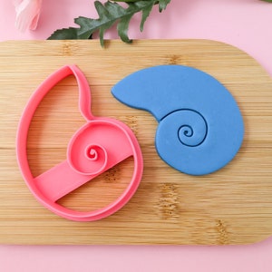 May include: A pink and blue set of cookie cutters shaped like seashells. The cutters are on a wooden surface.
