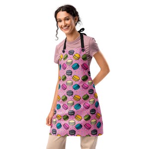 May include: A pink apron with a colorful macaron pattern. The apron features a repeating design of macarons in various colors, including yellow, blue, pink, green, and gray, with a sprinkle pattern. The apron has black straps and buttons.