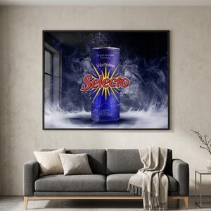 May include: A framed wall art print featuring a blue beverage can with the red text Selecto and a yellow starburst graphic, surrounded by swirling white smoke and mist effects against a dark blue background, displayed above a gray sofa.