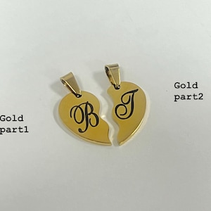 May include: Two gold-colored heart-shaped pendants, each with a letter engraved in black. One pendant has the letter "B", and the other has the letter "J". The pendants are designed to fit together, symbolizing a bond. The background is a plain, light color.