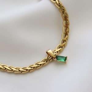 May include: Gold-toned necklace with a braided chain design. A rectangular, emerald-green gemstone pendant hangs from the chain. The necklace is displayed on a white, textured surface.