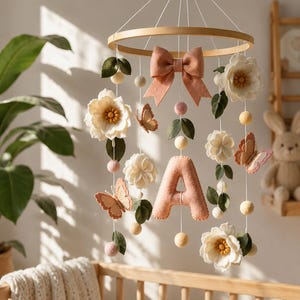 May include: A hanging mobile featuring a circular wooden frame with suspended felt elements including white flowers with yellow centres, peach-coloured butterflies, green leaves, small round felt balls in cream and pink, a large peach bow, and a letter A.
