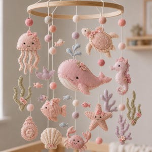 May include: A hanging mobile featuring felt sea creatures including a whale, turtle, seahorse, jellyfish, starfish, crab, fish, and seashells in shades of pink, lavender, and sage green, suspended from a circular wooden frame with felt balls.