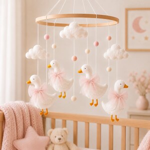 May include: A nursery mobile featuring a circular wooden frame with white felt clouds, four white felt ducks wearing soft pink tulle bows, and small felt pom-poms in shades of pink and cream suspended by white strings from the wooden ring.