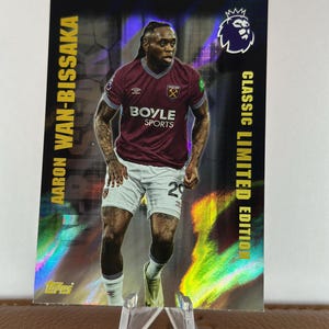Puede incluir: Tarjeta coleccionable con Aaron Wan-Bissaka en uniforme de fútbol granate y blanco. La tarjeta tiene el texto "Aaron Wan-Bissaka" y "Classic Limited Edition". Se muestra en un soporte de plástico transparente.