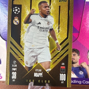 Puede incluir: Una tarjeta coleccionable Topps con Kylian Mbappé con una camiseta blanca con el logo de Emirates. La tarjeta tiene un diseño dorado y negro con el texto "100 Club" y otros detalles. La tarjeta está sobre un soporte de plástico transparente.