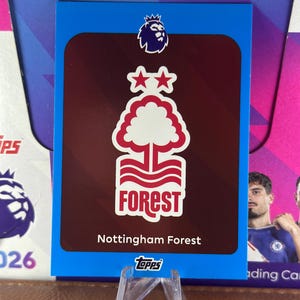 May include: A trading card featuring the Nottingham Forest logo, a white tree design with the word "FOREST" in white, and two stars on a maroon background. The card has a blue border and the Topps logo.