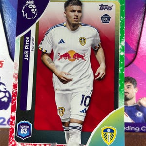 Pode incluir: Um cartão colecionável Topps com o jogador de futebol Joël Piroe com o uniforme branco e azul do Leeds United. O cartão tem um fundo vermelho e inclui o logotipo da Premier League, o logotipo da Topps e uma classificação de potência de 83.