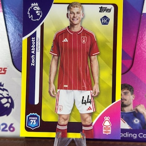 May include: A Zach Abbott trading card featuring the player in a red and white soccer uniform. The card has a yellow border and includes the Premier League logo, the Topps logo, and the text "Defender". The card is propped up on a clear plastic stand.