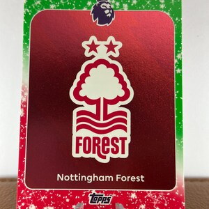 May include: A trading card featuring the Nottingham Forest logo, a white tree design with the word "FOREST" in white text. The card has a red background and is framed with a green border with white star accents. The Premier League logo is at the top.