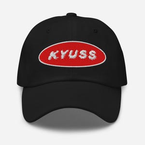 May include: Black baseball cap with a red oval patch. The patch features the embroidered word "KYUSS" in white, outlined in white. The cap has a curved brim and a button on top.