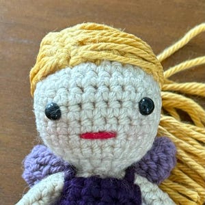 May include: A handmade crochet doll with a cream-colored face, black button eyes, and a red stitched mouth. The doll has yellow yarn hair and wears a purple dress with matching wings. The doll is a whimsical, handcrafted item.