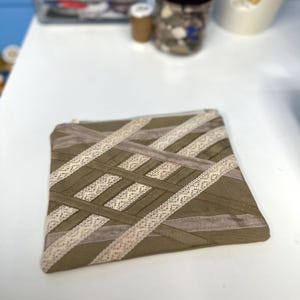 May include: A rectangular olive green fabric pouch featuring a decorative lattice pattern created with intersecting strips of cream-colored lace and smooth, matching olive green ribbon. The pouch includes a top zipper closure along the edge.