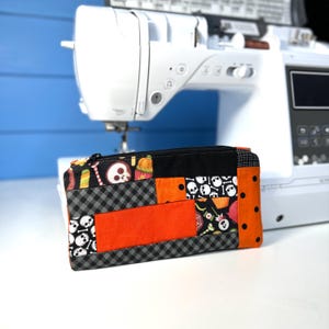 May include: A rectangular fabric zipper pouch featuring a patchwork design with orange, black, and white colors. The pattern includes skulls, candy, and pumpkins, combined with black and white gingham and solid orange fabric panels with black dots.