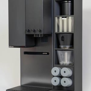 以下が含まれることがあります： ダークグレーのコーヒーマシン。収納ユニットが内蔵されています。ドリップトレイとアクセサリー用の複数のコンパートメントを備えた、洗練されたモダンなデザインです。「xbloom」のブランド名が前面に表示されています。