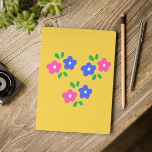 May include: A yellow notebook featuring a central graphic pattern of three clusters of small, five-petaled flowers in shades of pink and blue, each accompanied by small green leaves and a white center dot, arranged in a triangular formation.