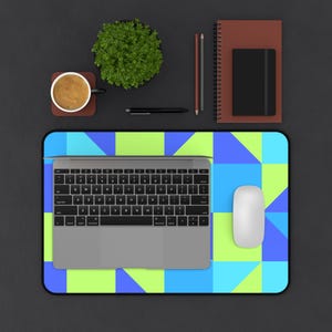 Op de afbeelding: Een rechthoekige bureaumat met een geometrisch patroon van limoengroene, felblauwe en cyaan vierkanten en driehoeken. Een zilveren laptop staat op de mat, vergezeld door een witte computermuis, een bruin notitieboek, een zwart dagboek en een koffiemok.