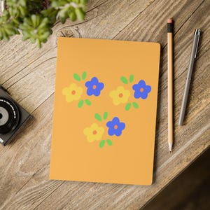 May include: A rectangular notebook with a solid orange cover featuring a repeating pattern of three clusters of small, five-petaled yellow and blue flowers with light green leaves. The notebook is positioned on a wooden surface with a pencil and pen.