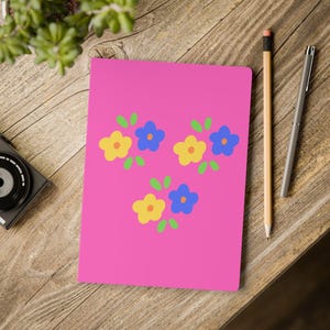 May include: A rectangular notebook with a vibrant pink cover featuring three clusters of stylized flowers. Each cluster consists of one yellow flower and one blue flower, accented by small green leaves. The notebook is positioned on a wooden surface.