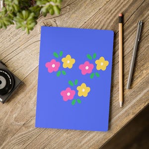 May include: A rectangular notebook with a solid blue cover featuring a repeating pattern of three clusters of small flowers. Each cluster consists of one pink flower and one yellow flower, both with white centers, surrounded by small green leaves.