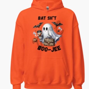 May include: Bright orange hoodie with a Halloween design. The graphic shows a cartoon ghost with a coffee cup and a leopard print bag, accompanied by bats and the text "BAT SH*T BOO-JEE".