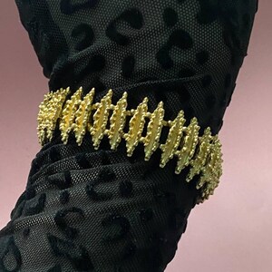 Pode incluir: Uma pulseira dourada com um padrão repetitivo de barras verticais e pequenos detalhes arredondados. A pulseira é usada sobre um tecido preto estampado com um desenho de leopardo. O fundo é rosa claro.