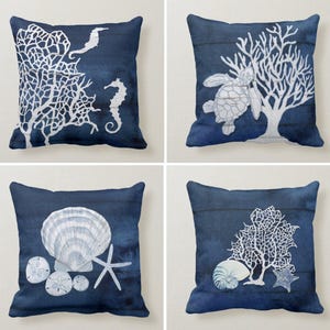 Pode incluir: Conjunto de quatro almofadas decorativas azul marinho com desenhos costeiros brancos. As almofadas apresentam corais, cavalos-marinhos, uma tartaruga marinha, conchas, estrelas do mar e um náutilo. Os desenhos são impressos em um fundo em aquarela.