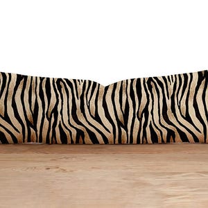 Peut inclure: Un long coussin rectangulaire avec un motif de rayures zébrées. Le coussin présente des rayures noires et beiges, créant un motif animalier audacieux. Le coussin est posé sur une surface en bois.