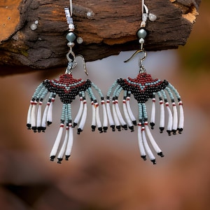 May include: A pair of handmade beaded earrings. The earrings feature a black, red, and light blue beaded design with white fringe. The earrings are suspended from silver-colored ear wires and are displayed against a blurred background.
