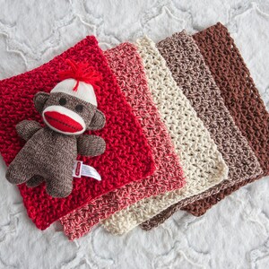 May include: A set of five crocheted baby blankets in red, pink, cream, brown, and beige. The blankets are arranged in a stack, with the red blanket on top. A small sock monkey toy is lying on the red blanket.