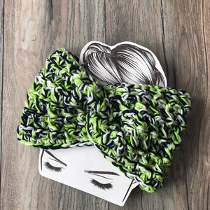 May include: A handmade, knitted headband in a vibrant mix of green, navy, and white yarn. The headband features a knotted bow design. The background is a wooden surface.