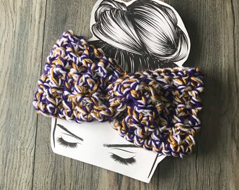 May include: A purple, gold, and white crocheted headband. The headband is in a knotted style and is shown on a white background.