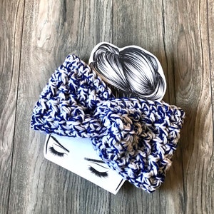 May include: A white and blue crocheted headband with a chunky knit texture. The headband is folded in half and has a bow shape.