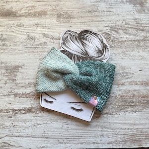 May include: A green and white knitted headband with a knotted bow. The headband is on a white wooden surface.