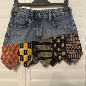 May include: A denim mini skirt with a patchwork design made from various patterned neckties. The skirt is hanging on a black hanger against a white background. The neckties feature red, yellow, black, and navy blue patterns.