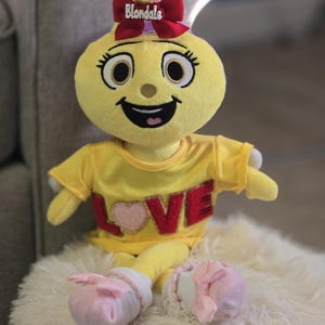 May include: A plush yellow doll with a smiling face, wearing a yellow shirt with the word "LOVE" and a pink heart. The doll has pink bows and the name "Blondale" on its head. It is sitting on a fluffy white surface.