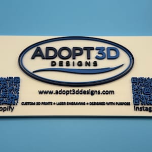 May include: A rectangular sign with the text "ADOPT3D DESIGNS" in blue and black, with a blue oval border. The sign also includes a website address, QR codes, and the words "Shopify" and "Instagram".