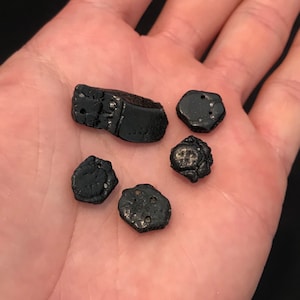 May include: A collection of small, irregularly shaped, black objects. The objects have a metallic sheen and varying textures, some with small holes or indentations. One object is rectangular, while the others are more rounded.