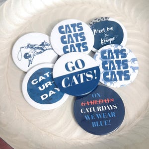 May include: A collection of seven circular pin-back buttons featuring various blue and white designs. Text includes "CATS CATS CATS," "meet me @ Kroger," "CAT-UR-DAY," "GO CATS!," and "ON GAMEDAYS CATURDAYS WE WEAR BLUE!" One button features a blue illustration of a cowboy.