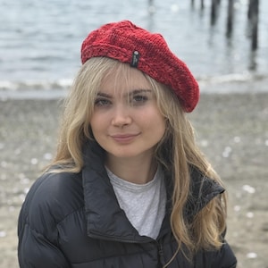 May include: A red knitted beret with a small tag on the side. The beret is worn by a person wearing a black puffer jacket and a gray shirt. The background shows a body of water and a pier.