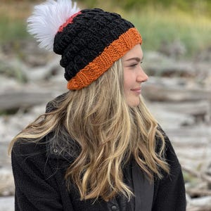 Op de afbeelding: Een zwarte gebreide muts met een oranje rand en een witte pompon. De hoed heeft een textuurpatroon en een rode streep boven de oranje rand. De hoed wordt gedragen door een persoon met lang blond haar.