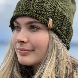 May include: A knitted olive green beanie with a ribbed design. A small leather tag with the text "Vashon Island Knits" is attached to the side of the hat. The hat is made of a thick, textured yarn.