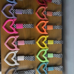 May include: A selection of heart-shaped accessories adorned with rhinestones, in shades of red, pink, orange, yellow, green, blue, purple, white, and silver. Each heart is connected to a black and white checkered band.