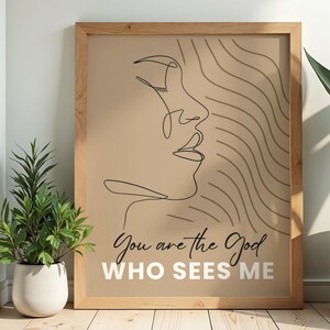 Puede incluir: Impresión artística enmarcada con un dibujo lineal minimalista de un rostro de perfil, con líneas onduladas en el fondo. El texto "You are the God WHO SEES ME" está impreso en una fuente elegante. El marco es de madera clara.
