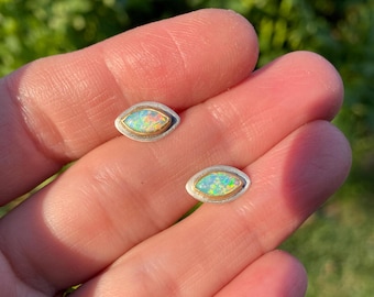 Puede incluir: Un par de pendientes de plata y oro con una pequeña piedra de ópalo iridiscente en cada uno. Los pendientes son de forma ovalada y tienen un acabado liso y pulido.