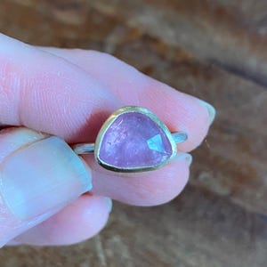 May include: A ring with a pink gemstone set in a gold bezel. The ring is on a finger.