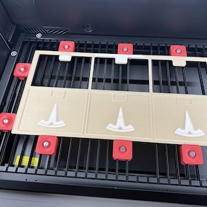 May include: A rectangular beige laser-cut template featuring three square sections, each embossed with a white Tesla logo. The template is secured onto a black metal honeycomb laser bed by eight red magnetic hold-down pins with silver centers.