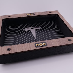 Puede incluir: Una bandeja de madera con interior negro y logotipo de Tesla. La bandeja tiene un marco de madera marrón claro con detalles en negro y herrajes decorativos dorados. El interior presenta un patrón de ondas en blanco y negro.